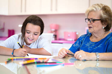 Smiling grandmother helping cheerful granddaughter drawing on paper while spending time together on kitchen at home - ADSF30686