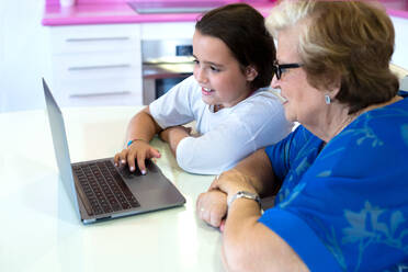 Seitenansicht einer lächelnden Enkelin und einer Großmutter, die am Tisch sitzen und einen Laptop in einem hellen Raum in einer Wohnung benutzen - ADSF30690