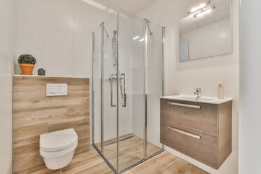 Glasduschkabine zwischen Toilette und Waschbecken unter einem an der gefliesten Wand hängenden Spiegel im Badezimmer - ADSF30725