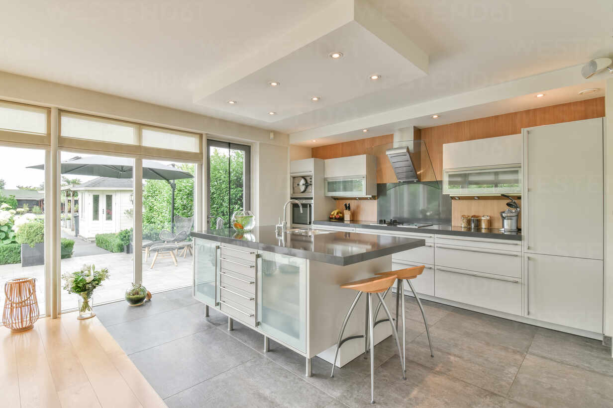 Zeitgenössische Küche Interieur mit Tisch und Hocker gegen Kühlschrank und  Schränke in Haus mit Glaswand in der Tageszeit, lizenzfreies Stockfoto