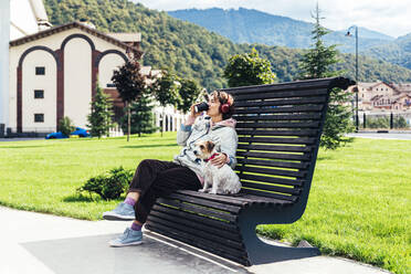 Frau trinkt Kaffee mit Hund auf einer Bank an einem sonnigen Tag - OMIF00099