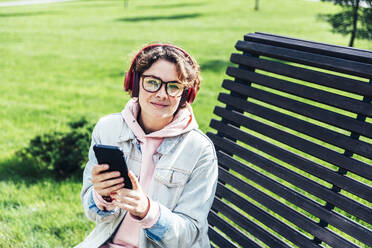 Lächelnde Frau mit Brille hält Handy auf Bank - OMIF00101