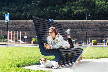 Frau benutzt Mobiltelefon, während sie im Park auf einer Bank neben einem Hund liegt - OMIF00103