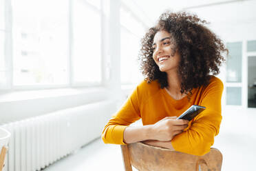 Lächelnde Afro-Frau, die ein Smartphone hält, während sie auf einem Stuhl zu Hause sitzt - KNSF09067