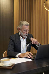 Mature businessman holding eyeglasses while working on laptop in hotel - VEGF05081