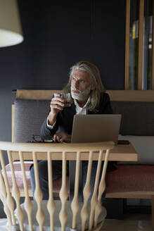 Nachdenklicher Mann mit Laptop, der ein Whiskey-Glas hält, während er im Hotel sitzt - VEGF05096