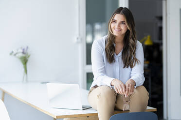 Smiling business professional sitting on desk at workplace - KNSF09125