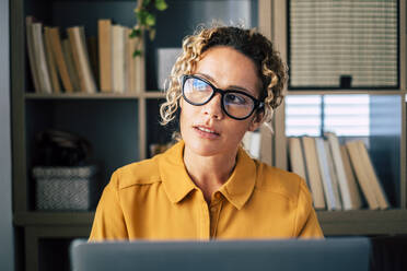 Thoughtful businesswoman wearing eyeglasses sitting with laptop at home - SIPF02482