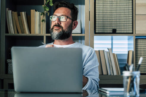 Nachdenklicher Geschäftsmann mit Laptop am Schreibtisch im Heimbüro - SIPF02487