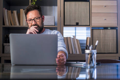Männlicher Berufstätiger mit Brille, der auf einen Laptop auf dem Schreibtisch im Heimbüro schaut - SIPF02492