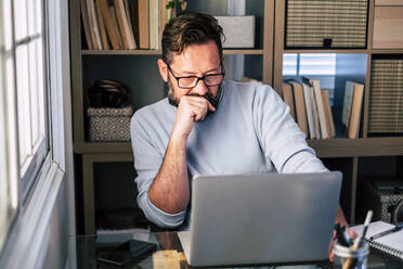 Männlicher Berufstätiger mit Hand am Kinn, der zu Hause am Laptop arbeitet - SIPF02497