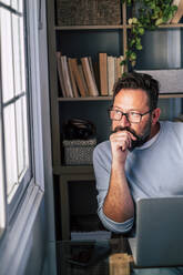 Thoughtful male freelancer looking through window while working at home - SIPF02498