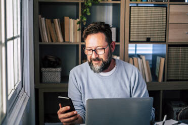 Geschäftsmann, der im Büro zu Hause sein Smartphone am Laptop benutzt - SIPF02500