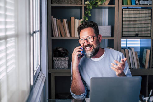 Männlicher Freiberufler gestikuliert, während er im Büro zu Hause mit seinem Smartphone telefoniert - SIPF02501