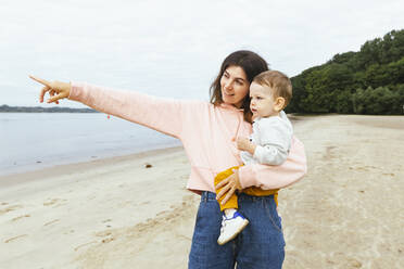 Mother carrying male toddler pointing while standing at beach - IHF00508