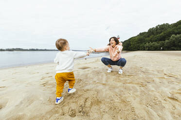 Männliches Kleinkind geht auf seine Mutter zu, die am Strand im Sand hockt - IHF00509