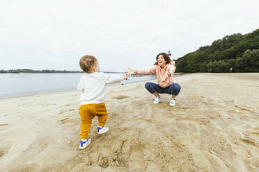 Male toddler walking towards mother crouching on sand at beach - IHF00509