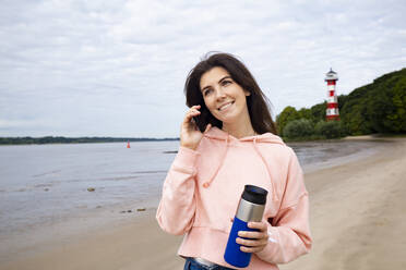 Lächelnde Frau, die mit ihrem Smartphone telefoniert und einen isolierten Getränkebehälter am Strand hält - IHF00515