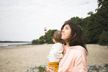 Frau schaut nach oben, während sie ihren Sohn am Strand trägt - IHF00518