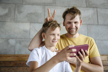 Lächelnder Mann gestikuliert, während sein Sohn auf einer Bank ein Selfie mit dem Handy macht - EYAF01783