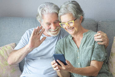 Älterer Mann winkt während eines Videoanrufs über sein Smartphone, während er neben einer Frau zu Hause sitzt - SIPF02511