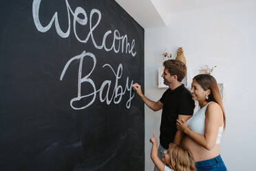 Male writing Welcome Baby inscription on blackboard against expectant female beloved and daughter in house - ADSF30807