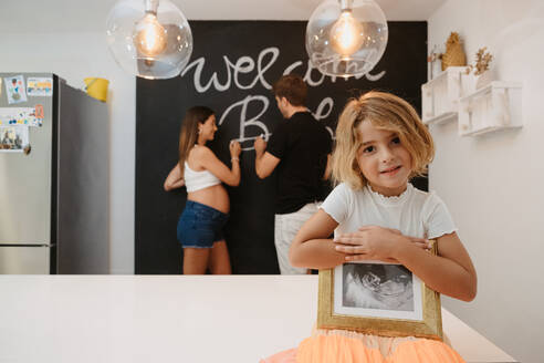 Charming child with future sibling ultrasound picture looking at camera against pregnant mother and father writing on chalkboard in house - ADSF30808