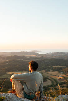 Back view of unrecognizable male traveler admiring ocean and plantations with trees while resting on ridge at sunset - ADSF30813