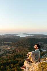 Seitenansicht männlicher Reisender, der das Meer und die Plantagen mit Bäumen bewundert, während er sich auf einem Bergrücken bei Sonnenuntergang ausruht - ADSF30814
