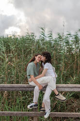 Side view of charming young homosexual girlfriends spending time on fence under cloudy sky in evening countryside - ADSF30816