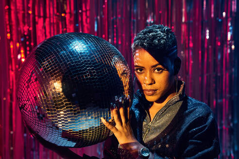 Trendy young ethnic female looking at camera while holding mirror ball in nightclub - ADSF30824