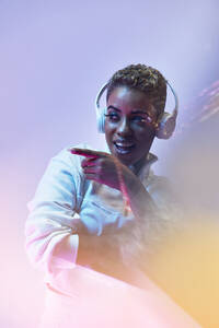 Neugierige junge trendige afroamerikanische Frau mit Make-up in drahtlosen Kopfhörern, die auf hellem Hintergrund wegschaut - ADSF30842