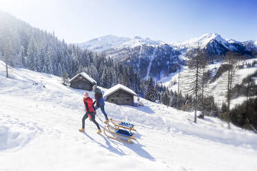 Paar, das einen Schlitten zieht, während es an einem sonnigen Tag im Schnee spazieren geht - HHF05731