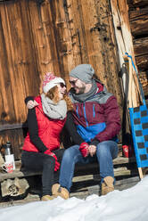 Mann mit Arm um Frau vor Hütte sitzend an einem sonnigen Tag im Winter - HHF05743