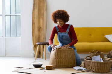 Junge Afro-Frau beim Reinigen eines Weidenkorbs im Wohnzimmer - GIOF13640