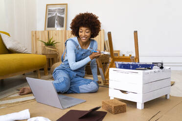Afro-Frau benutzt Laptop, während sie einen Stuhl im Wohnzimmer bemalt - GIOF13690