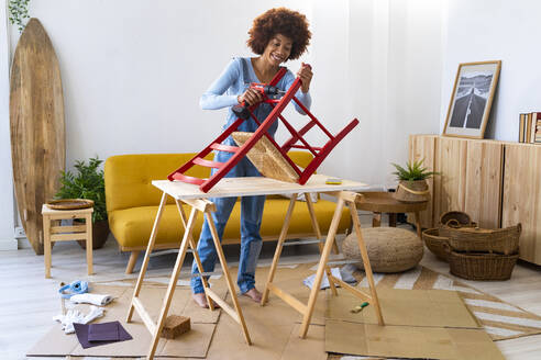 Young woman repairing chair with electric screwdriver in living room - GIOF13698