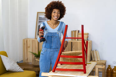 Smiling redhead woman with electric screwdriver standing at table - GIOF13699