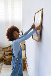 Happy woman adjusting picture frame on white wall - GIOF13711