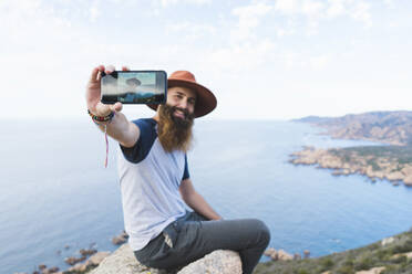 Bärtiger Mann nimmt Selfie durch Smartphone, während er auf einem Felsen sitzt - AFVF09232