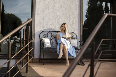 Junge Frau mit Buch auf einer Bank auf der Terrasse sitzend - LLUF00129