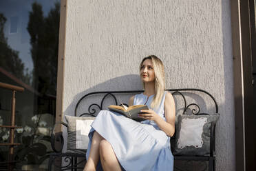 Nachdenkliche Frau mit Buch sitzt auf einer Bank auf der Terrasse an einem sonnigen Tag - LLUF00130