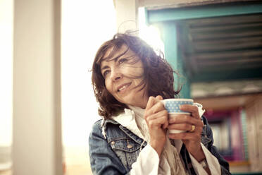 Smiling woman holding coffee mug in hut - AJOF01655