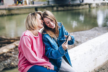 Smiling daughter using mobile phone while sitting with mother on retaining wall - OMIF00109