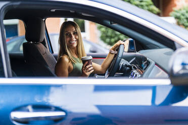 Smiling woman holding coffee cup while sitting in car - DLTSF02216