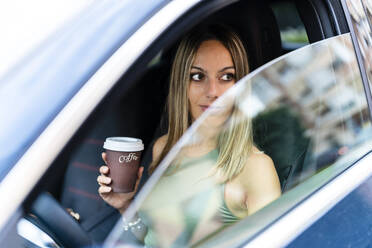 Blonde Frau mit Kaffeetasse schaut durch ein Autofenster - DLTSF02218