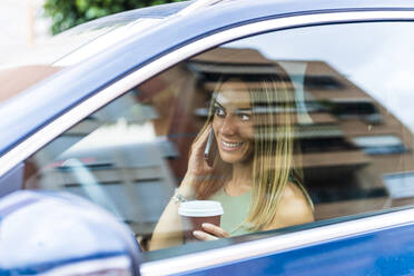 Lächelnde Frau mit Kaffeetasse, die im Auto mit ihrem Smartphone telefoniert - DLTSF02219