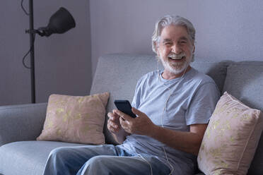 Happy man with mobile phone and in-ear headphones sitting at home - SIPF02527