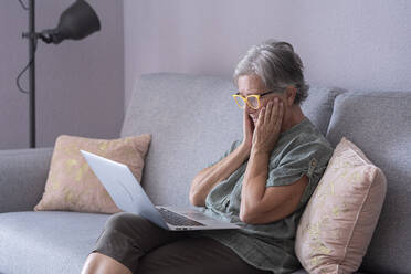 Besorgte Frau mit Laptop auf dem Sofa im Wohnzimmer sitzend - SIPF02530