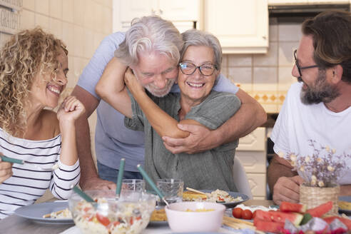 Ältere Frau, die einen Mann umarmt, während sie mit ihrer Familie am Esstisch sitzt - SIPF02535